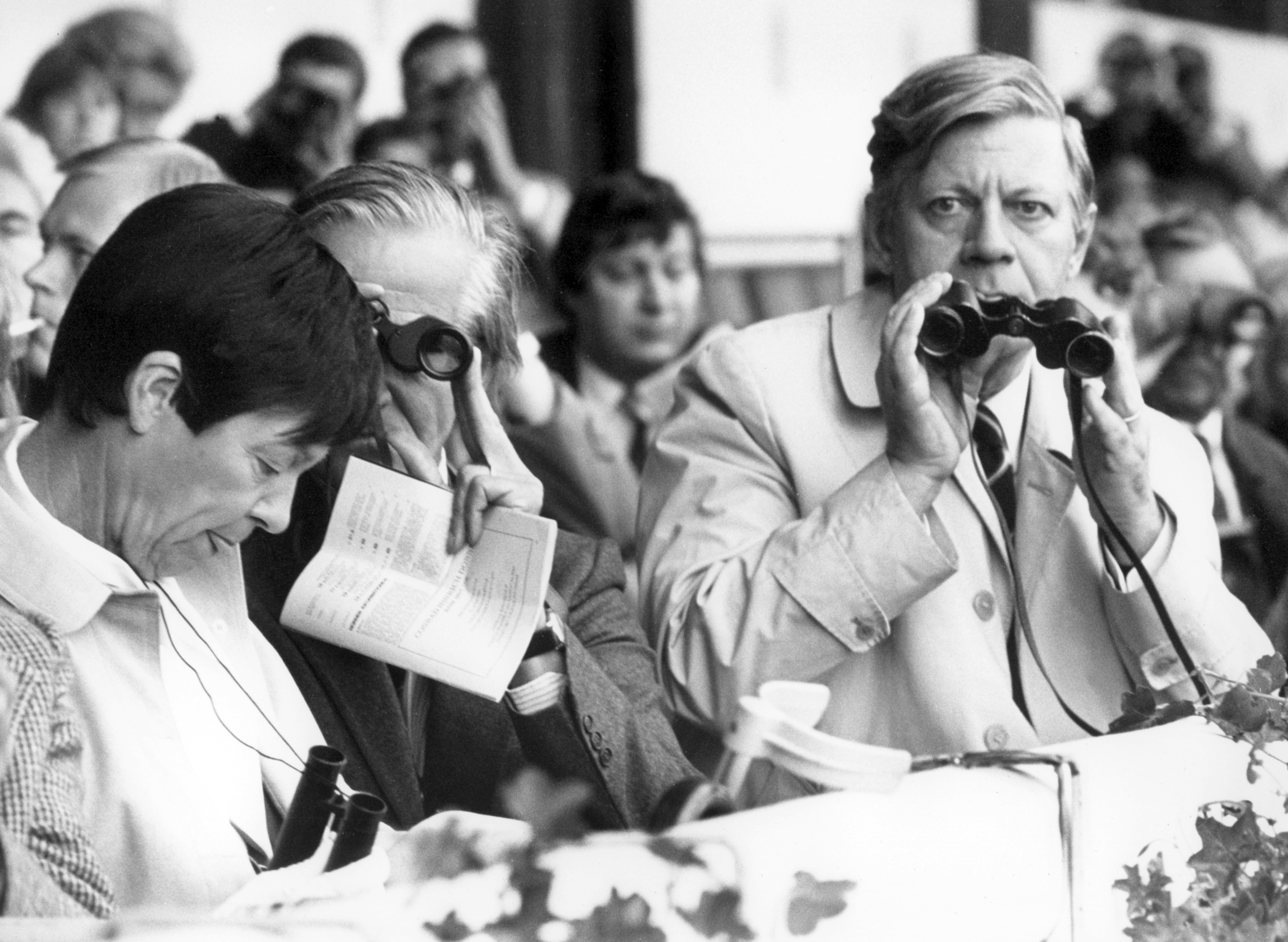 Helmut und Loki Schmidt auf der Tribüne beim Pferderennen. Er und ein weiterer Herr schauen durch Ferngläser in Richtung Bahn.
