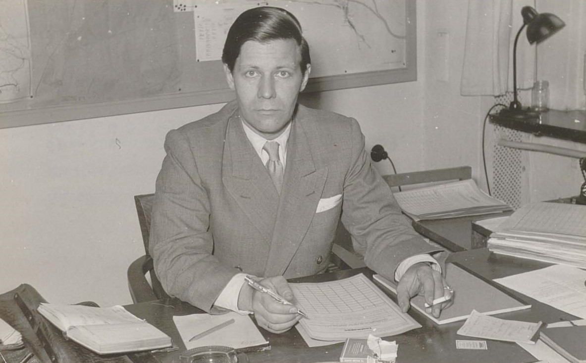 Helmut Schmidt sitzt an einem Schreibtisch in der Verkehrsbehörde und bearbeitet Dokumente. Er blickt ernst direkt in die Kamera.