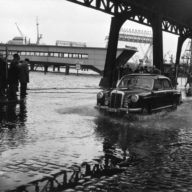 Ein schwarzer Mercedes fährt durch das stehende Wasser auf der Straße unterhalb des Hochbahn-Viadukts. An den Seiten stehen Menschen.